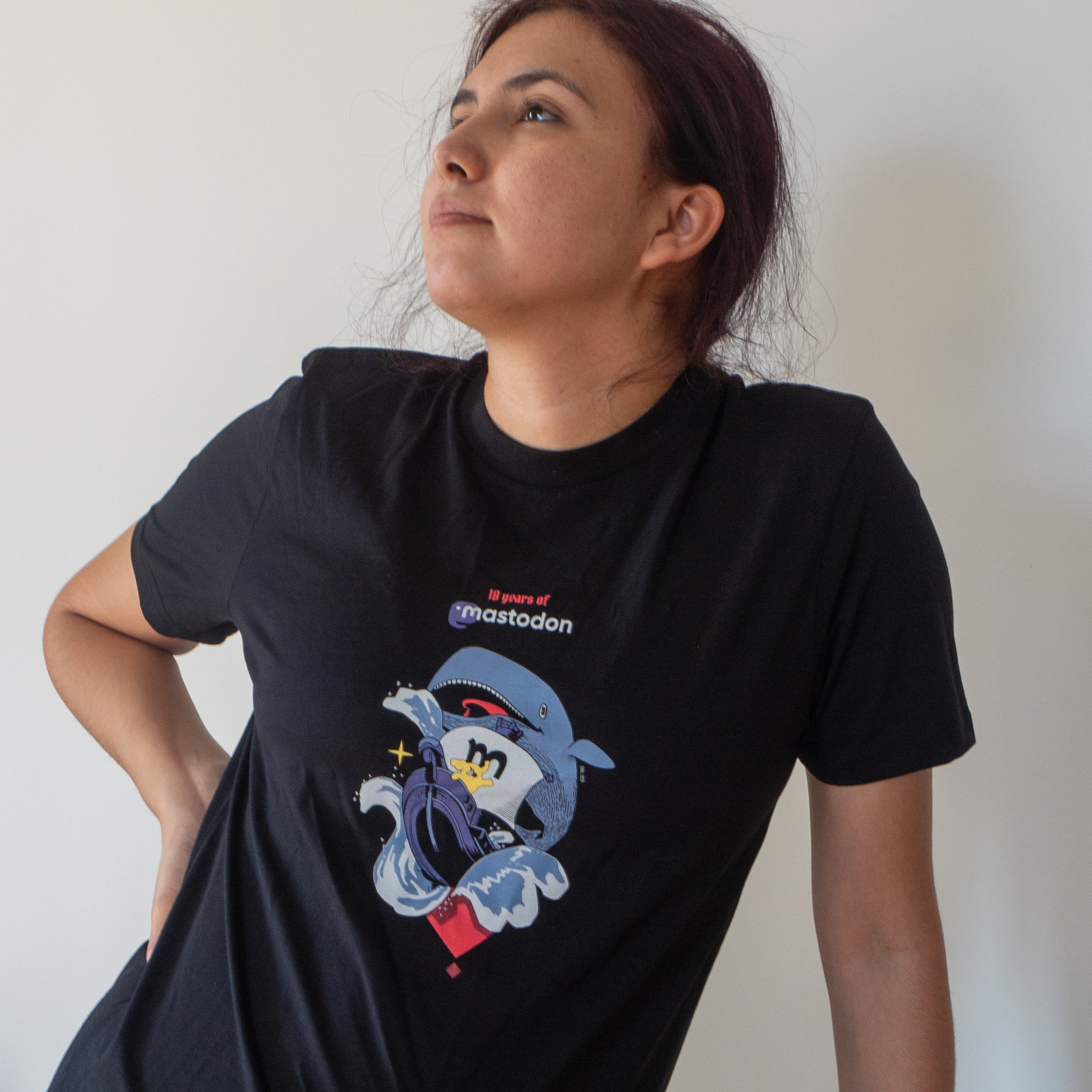 A beautiful woman demonstrating the Mastodon 10th anniversary t-shirt featuring a little Mastodon on a sailboat next to a big white whale under the caption 10 years of Mastodon.